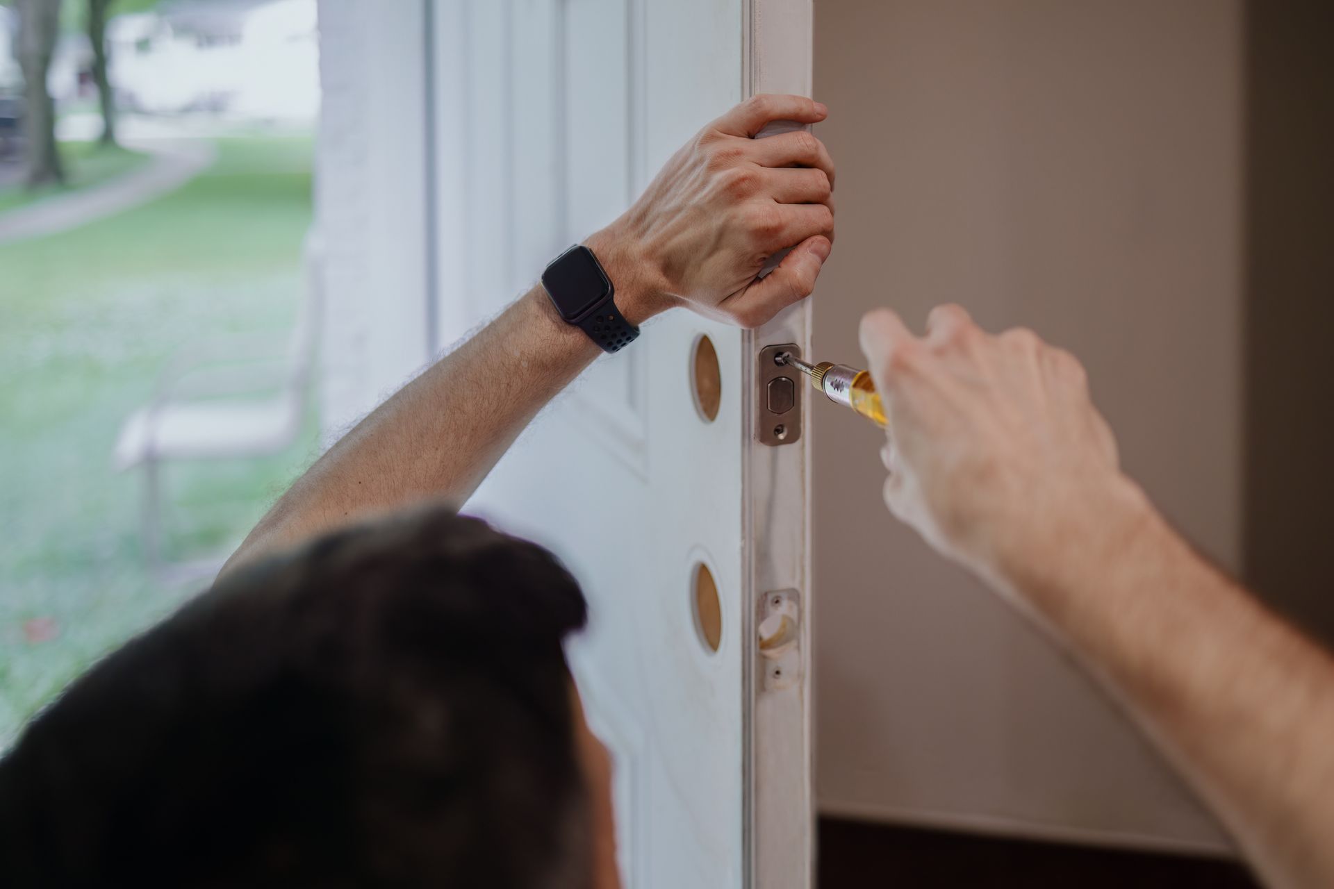 Hands using screwdriver to replace door lock with part of outdoor area visible behind.