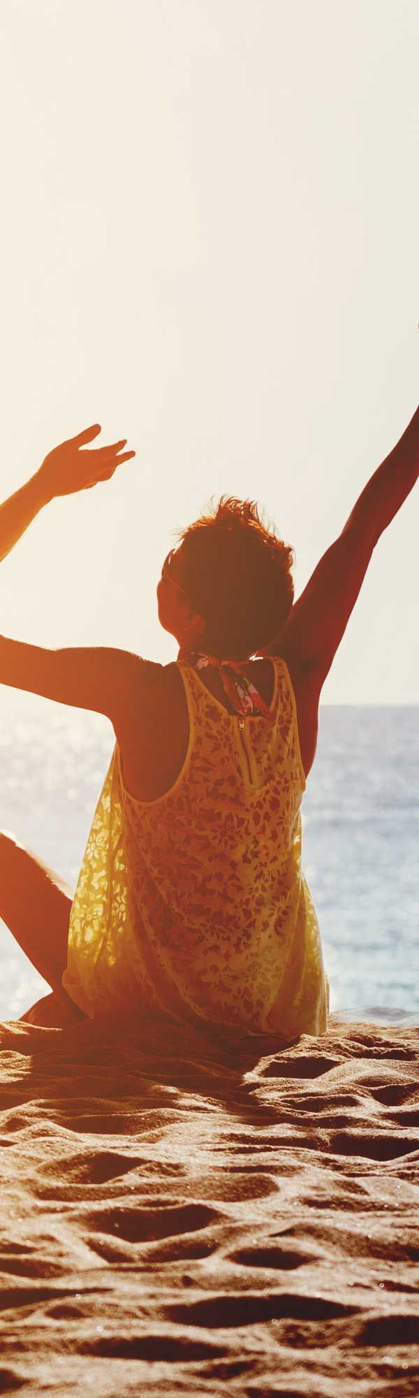 Person with arms raised towards the sun, sitting on a beach, overlooking the ocean.
