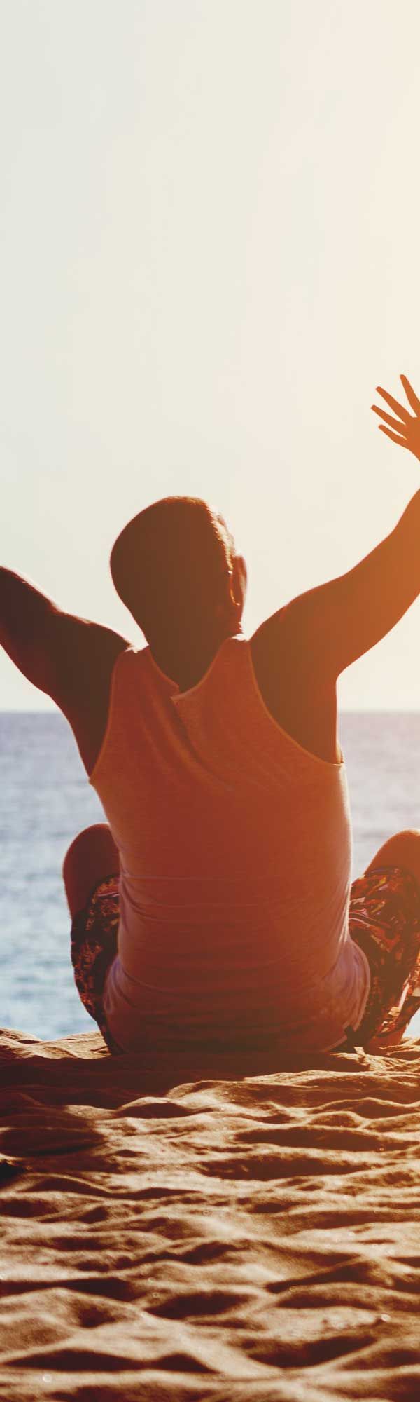 Person seated on beach with arms raised toward sunlight over the ocean.