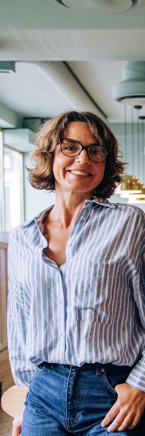 Woman in glasses smiling, wearing a blue striped shirt and jeans. Leaning against a table in a bright room.