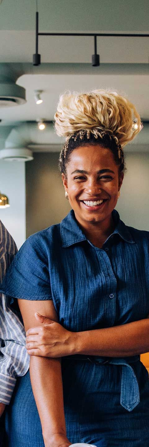 Woman smiles, arms crossed, wearing a denim dress, in an indoor setting. Her hair is in a bun.