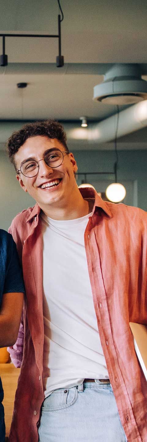 Man smiling, wearing glasses and a pink shirt over a white tee. In an office setting.