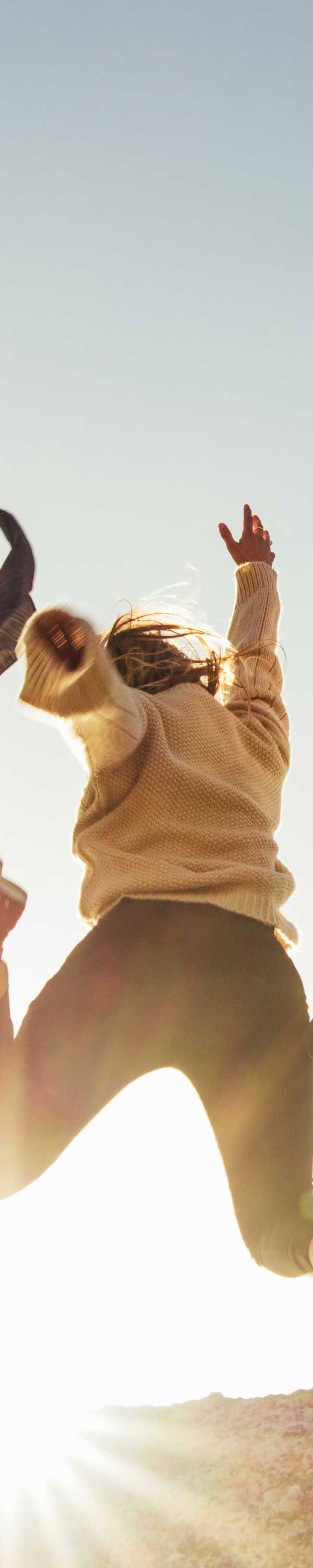 Person jumping in the air in front of a bright sun. They wear a light-colored sweater and dark pants.