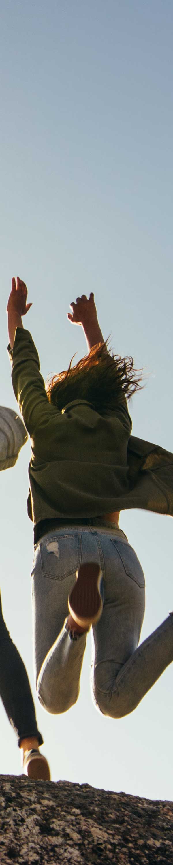 A person jumping in the air against a clear sky. They wear a green jacket and light-colored pants.