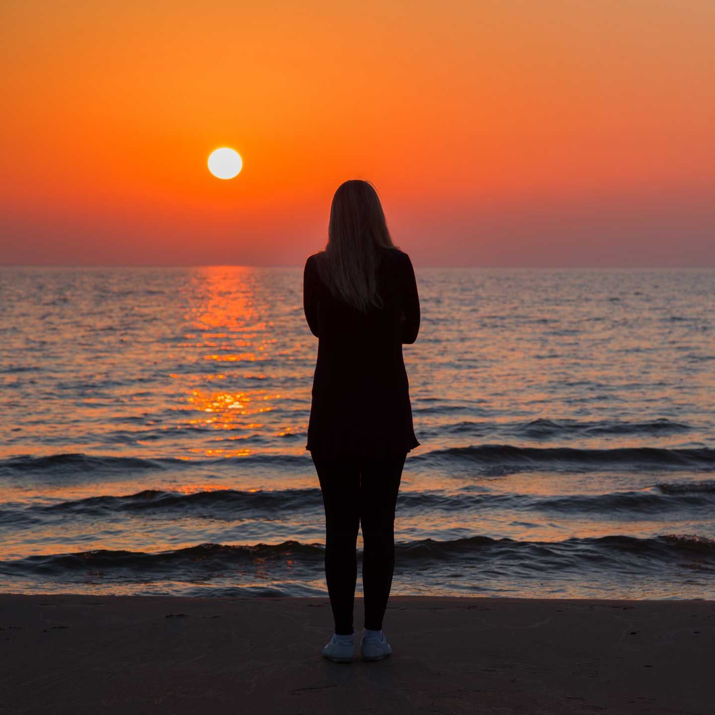 Woman standing on a beach, facing the setting sun over the ocean. Orange sky, dark silhouette. Woman standing on a beach, facing the setting sun over the ocean. Orange sky, dark silhouette.