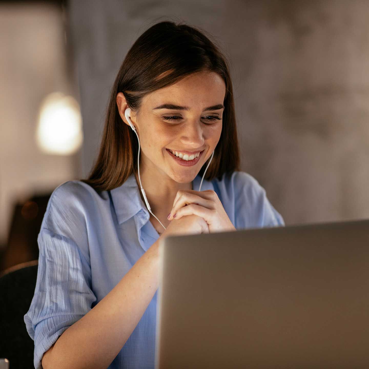 Woman smiling, wearing earbuds, looking at laptop screen in a dim room. Woman smiling, wearing earbuds, looking at laptop screen in a dim room.