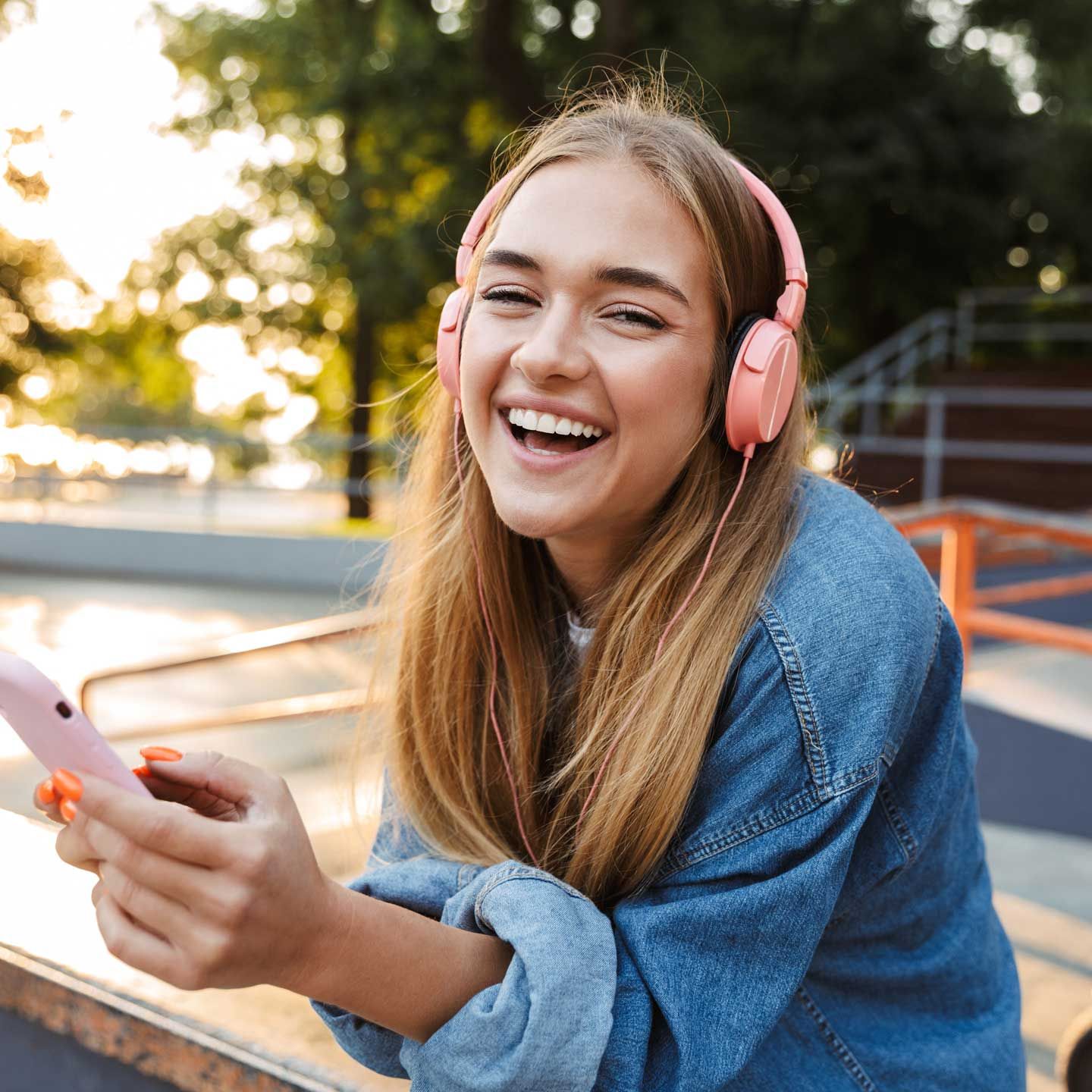 Woman with pink headphones and phone smiles, leaning on a skate park. Woman with pink headphones and phone smiles, leaning on a skate park.