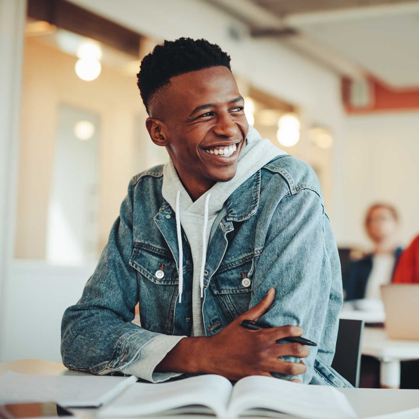 Young person smiles while sitting at a desk with a book in a classroom; wears a denim jacket and hoodie. Young person smiles while sitting at a desk with a book in a classroom; wears a denim jacket and hoodie.