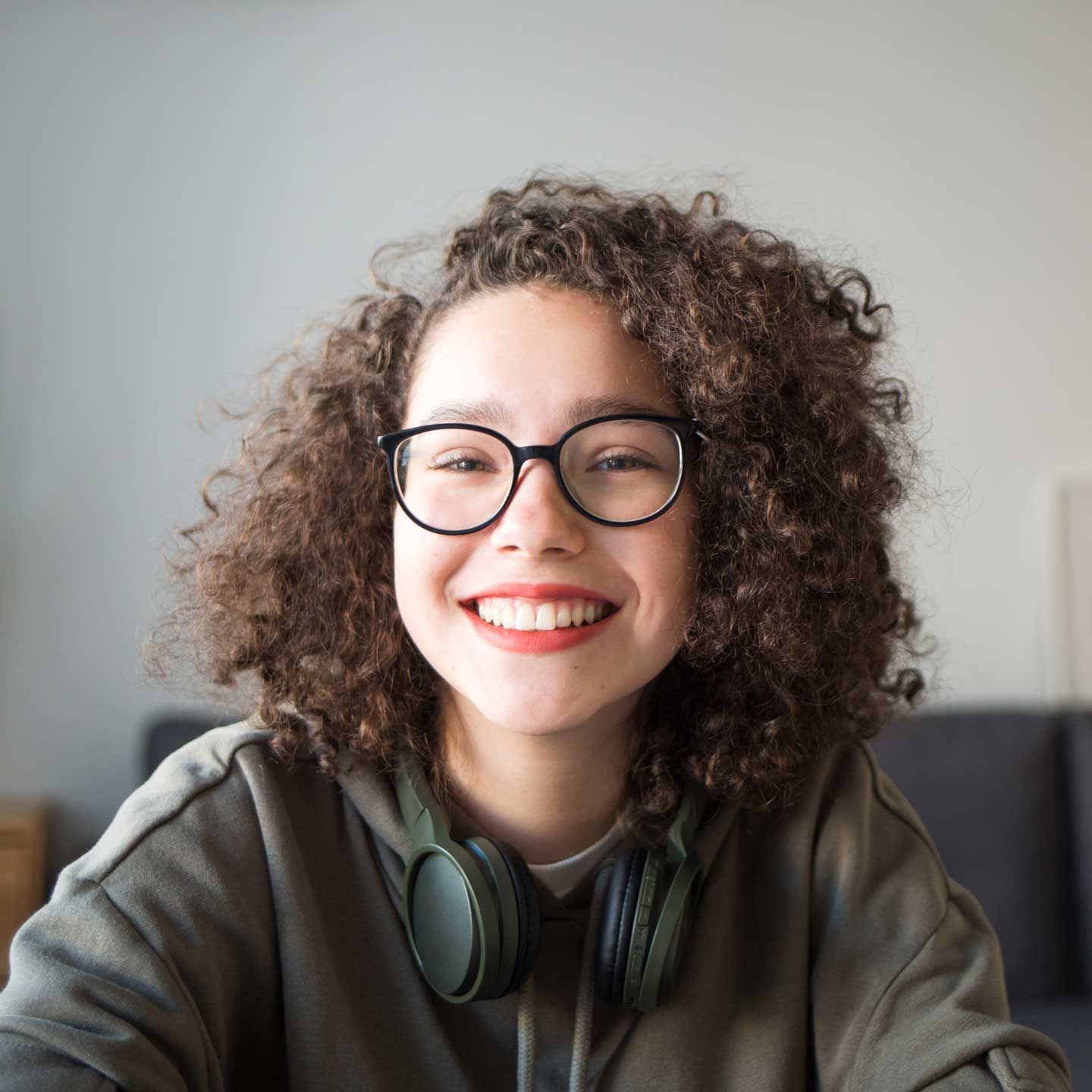 Woman with curly hair and glasses smiles at the camera, wearing headphones and a green hoodie. Woman with curly hair and glasses smiles at the camera, wearing headphones and a green hoodie.