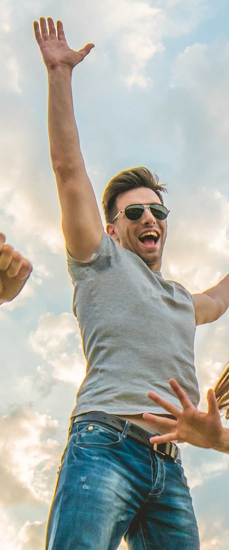 A man in sunglasses jumping with arms raised, smiling against a cloudy sky.