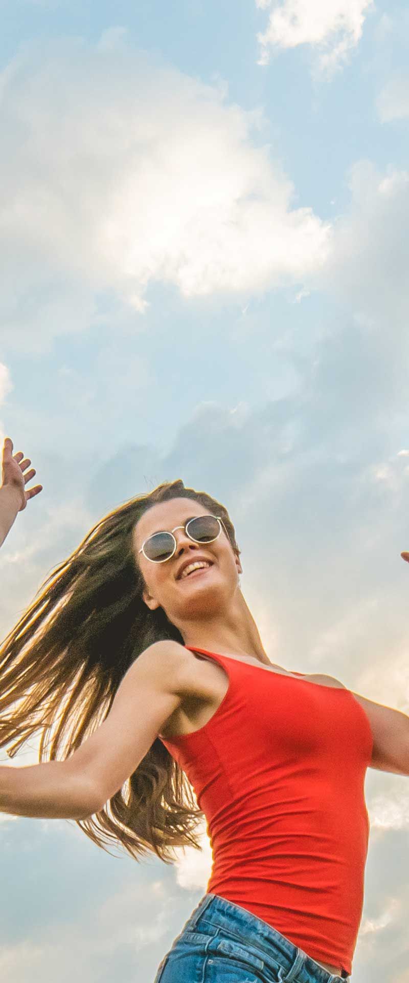 Woman in red top and sunglasses jumps with joy against a cloudy sky.