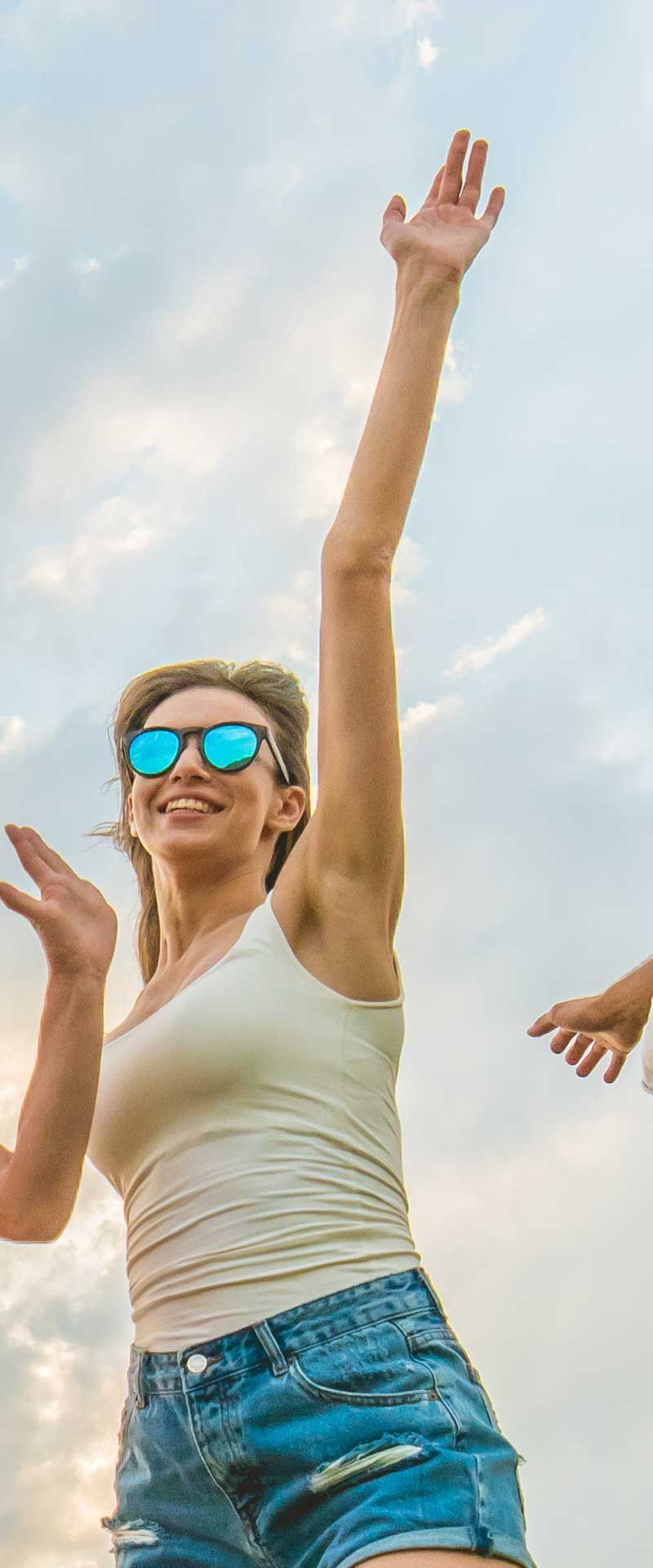 Woman in sunglasses, tank top, and shorts, arms raised in front of a cloudy sky.