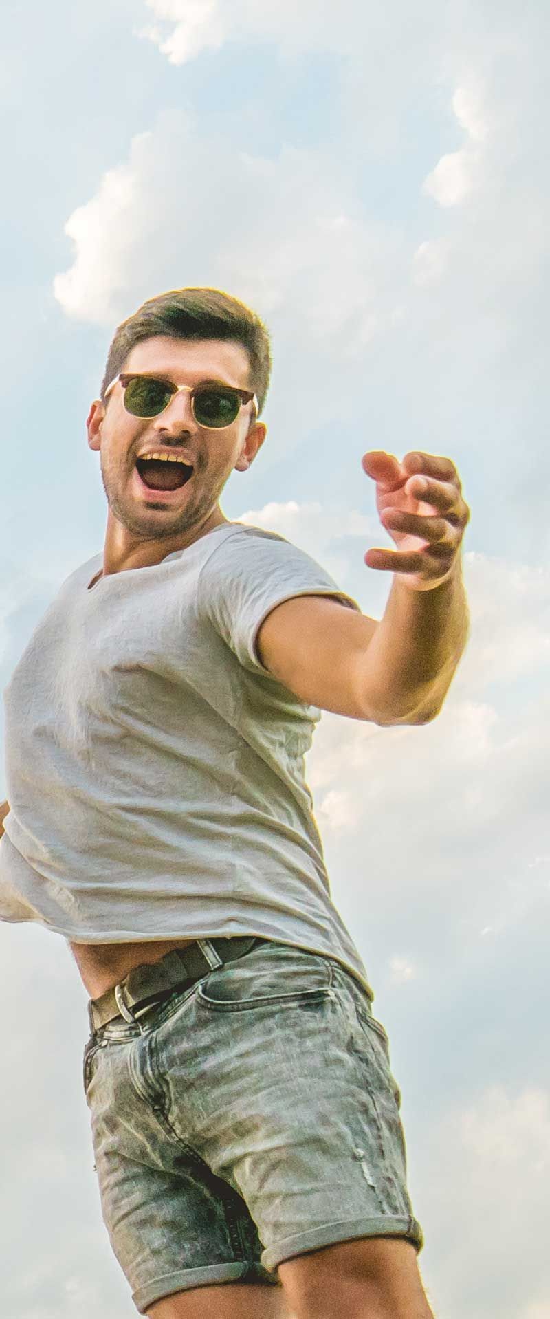 Man wearing sunglasses, grey shirt and shorts, jumping with arms outstretched against a cloudy sky.