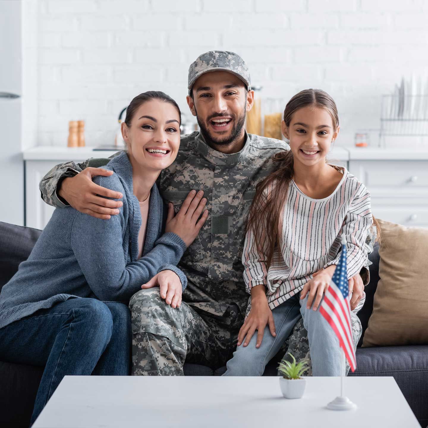 Family in living room: soldier in camo uniform smiles with wife, daughter; American flag on table. Family in living room: soldier in camo uniform smiles with wife, daughter; American flag on table.