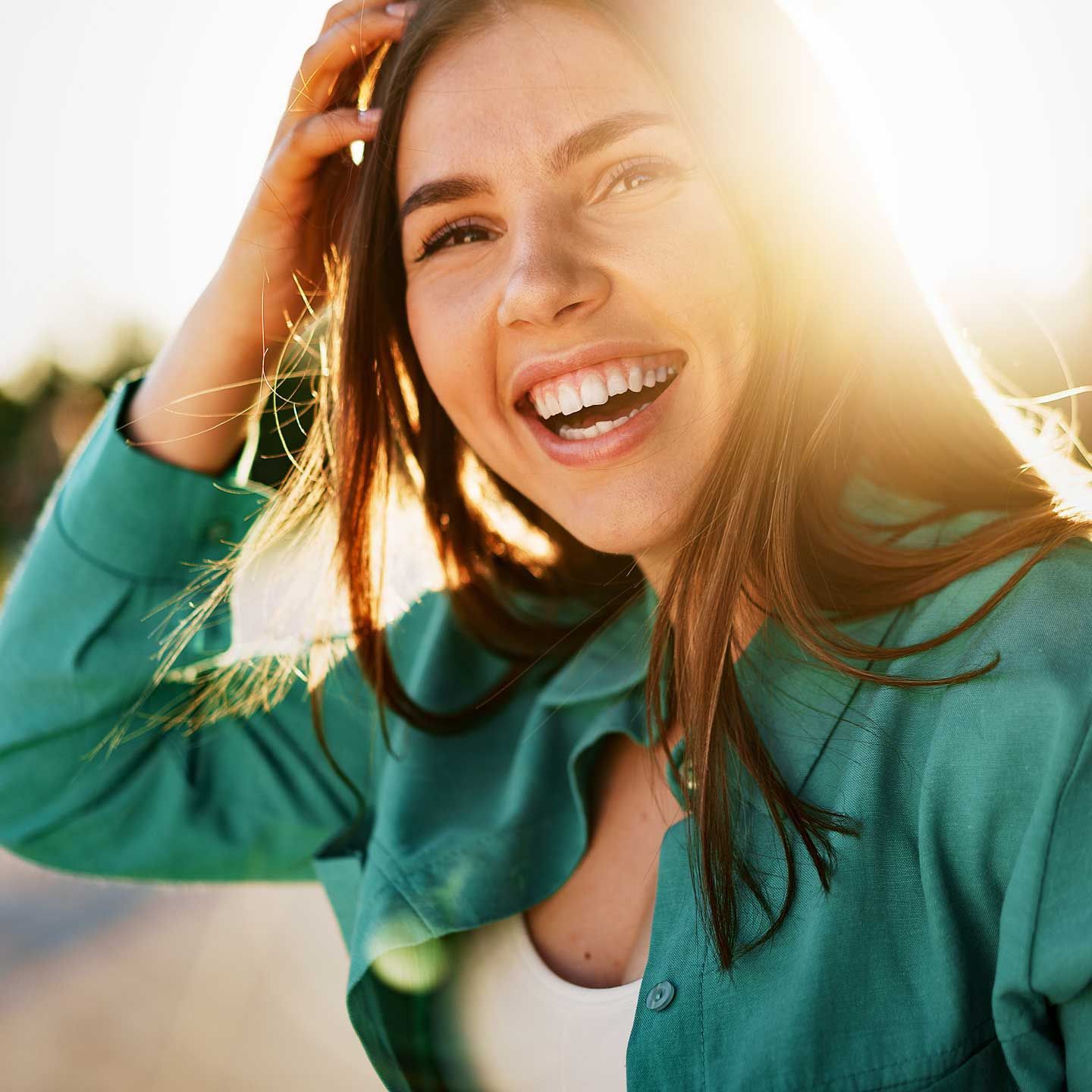 Woman with long brown hair, smiling widely, touches her head, sunlit background. Woman with long brown hair, smiling widely, touches her head, sunlit background.