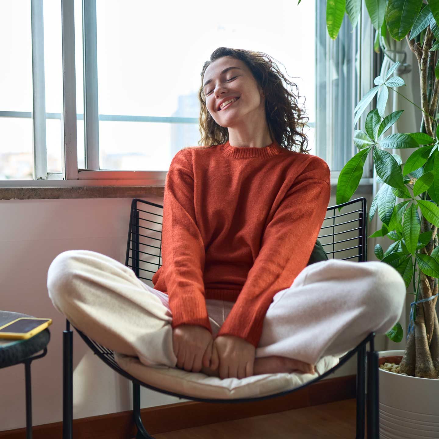 Woman in orange sweater and white pants smiles, sitting in chair by a window, near a potted plant. Woman in orange sweater and white pants smiles, sitting in chair by a window, near a potted plant.