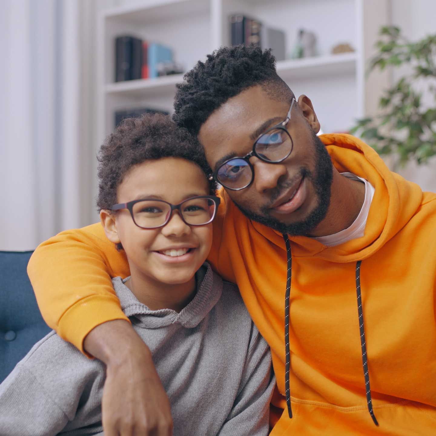 Man in orange hoodie with arm around a smiling child wearing glasses, indoors. Man in orange hoodie with arm around a smiling child wearing glasses, indoors.