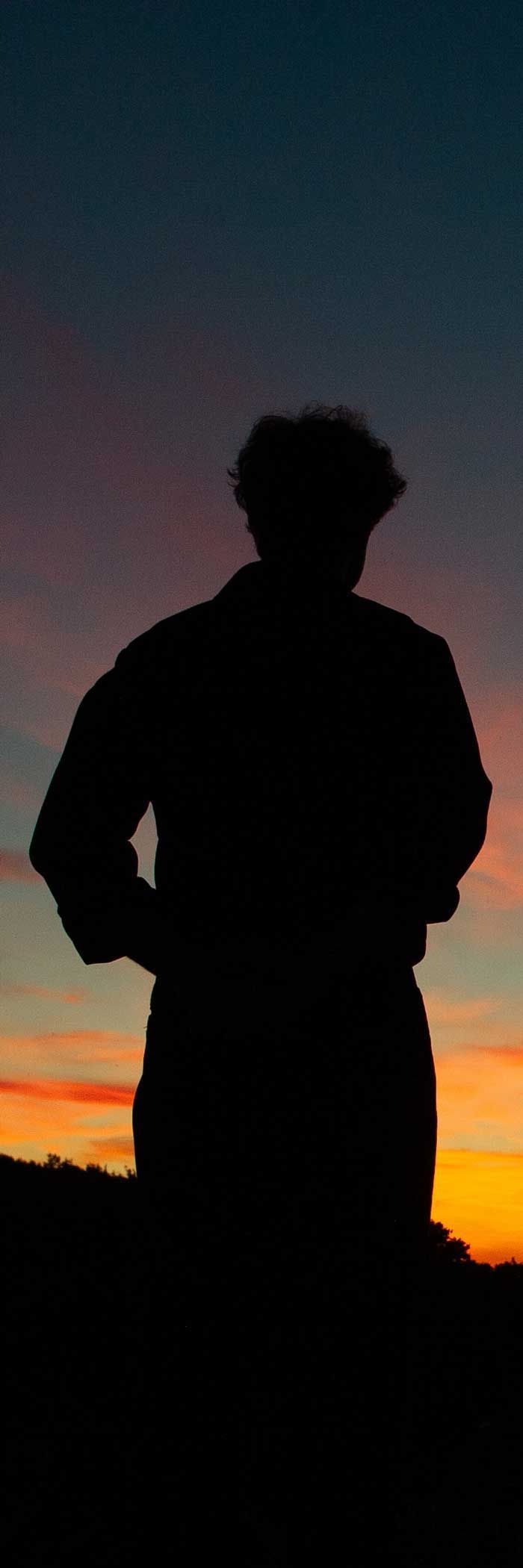 Silhouette of a person standing at sunset, hands on hips, against a colorful sky.