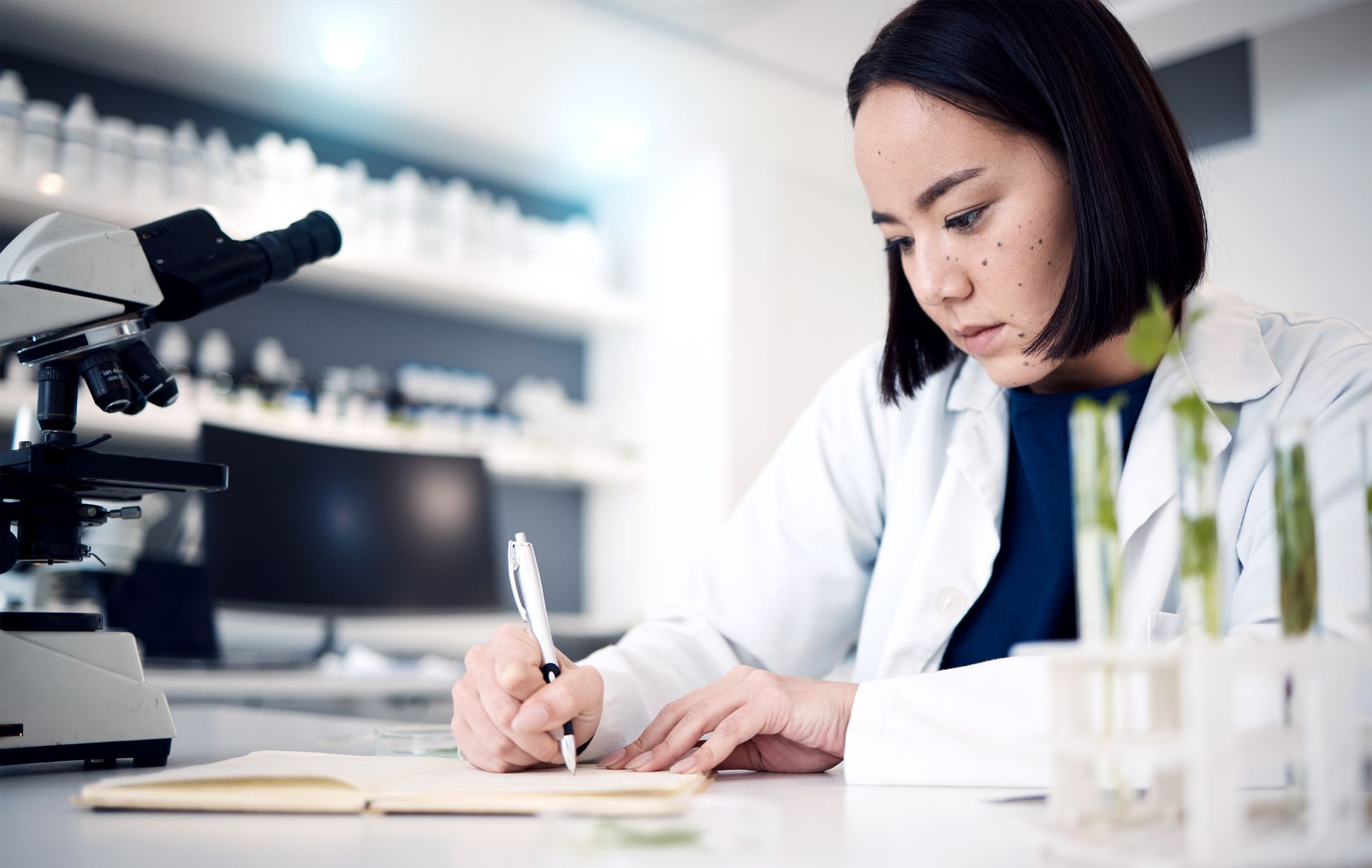 Woman in lab coat writing notes in her notebook whilst in the lab