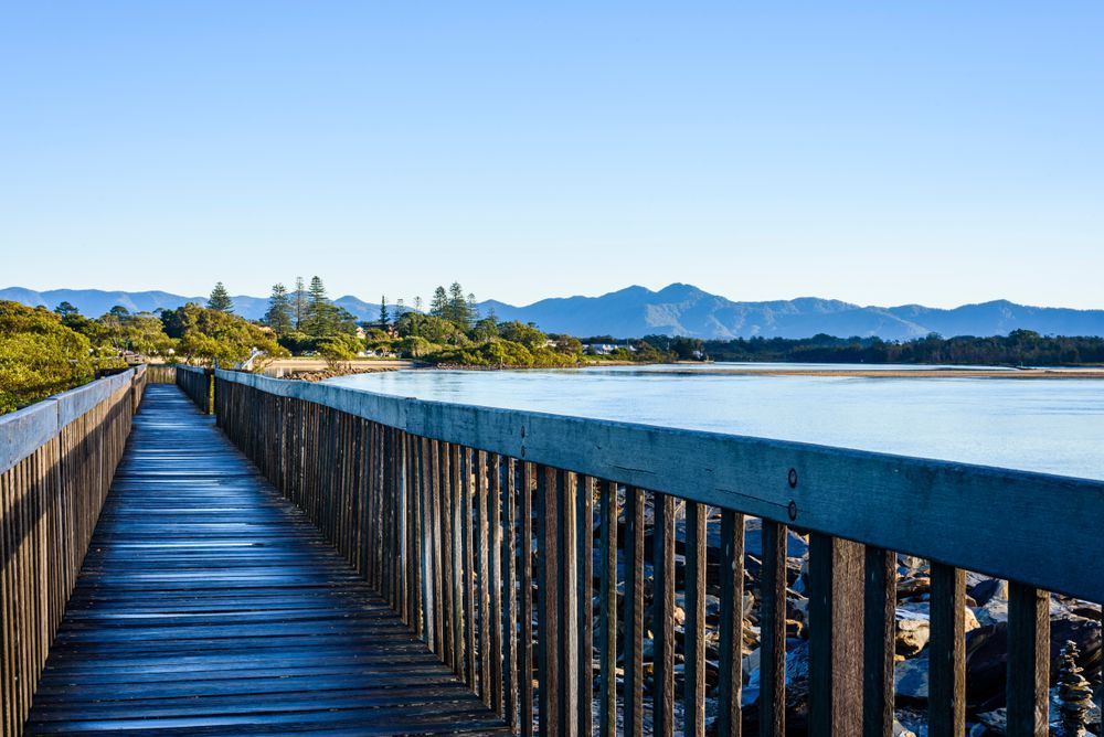 A Wooden Bridge Over A Body Of Water With Mountains In The Background — Boambee Home Improvements in Urunga, NSW