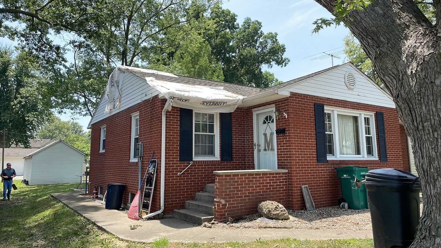 A small brick house with black shutters and a tree in front of it.