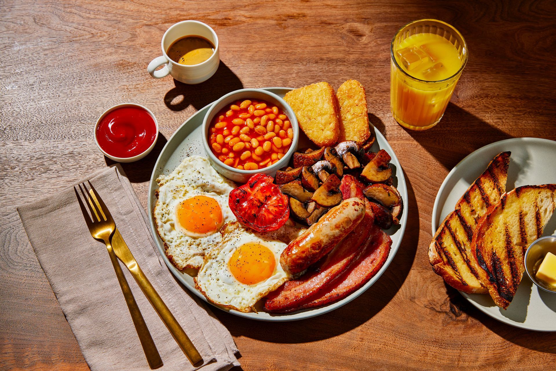 A plate of food with eggs , sausages , beans , hash browns and toast on a wooden table.