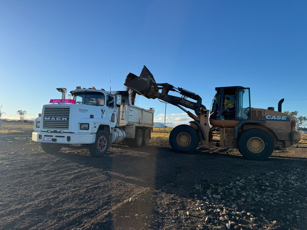 A front-end loader dumping gravel into a white Mack truck on a gravel lot under a blue sky.