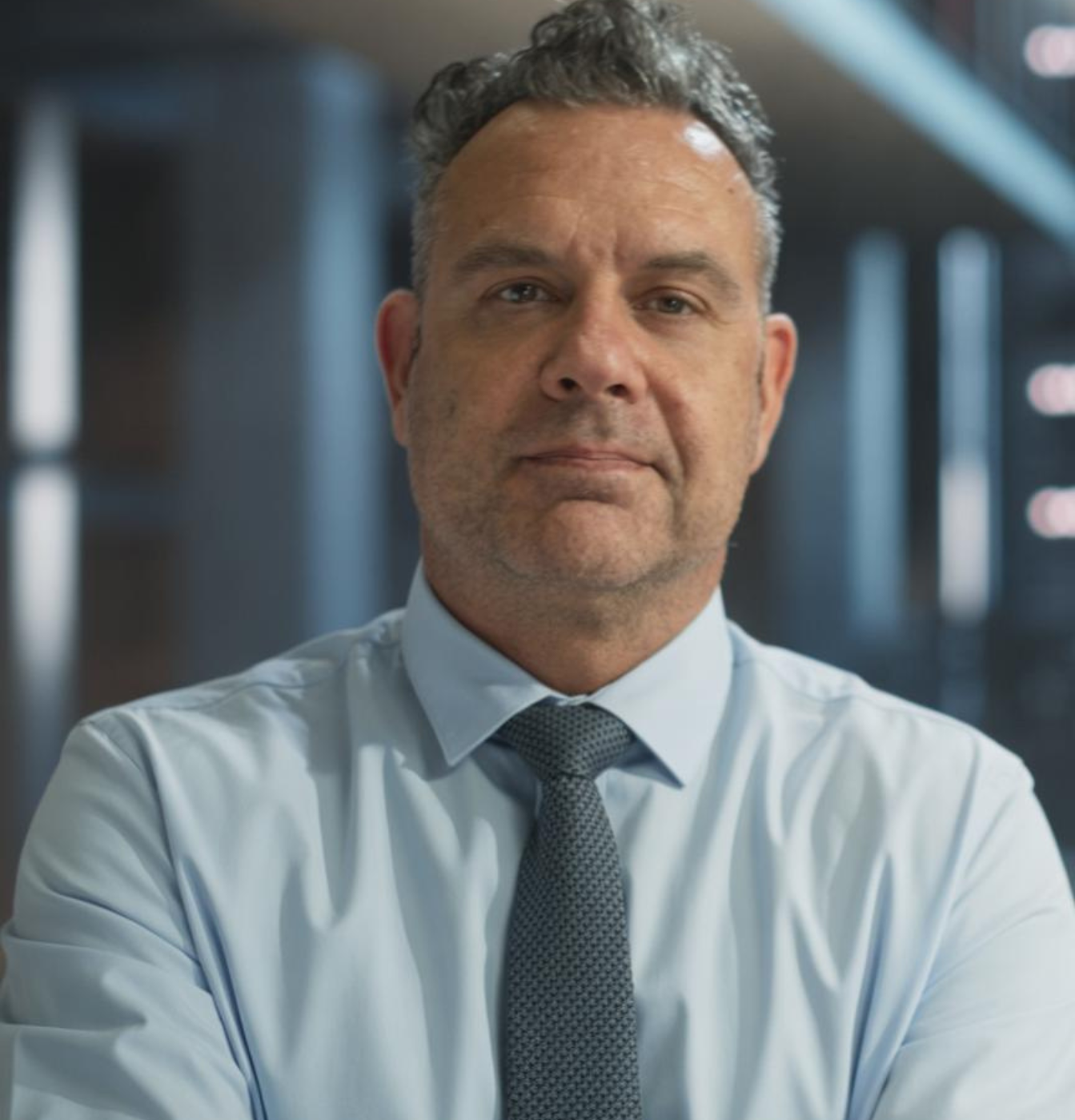 Man in light blue shirt and tie, looking forward, in office setting.