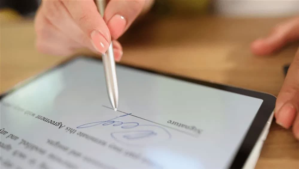 A hand signing a document on a tablet with a stylus.