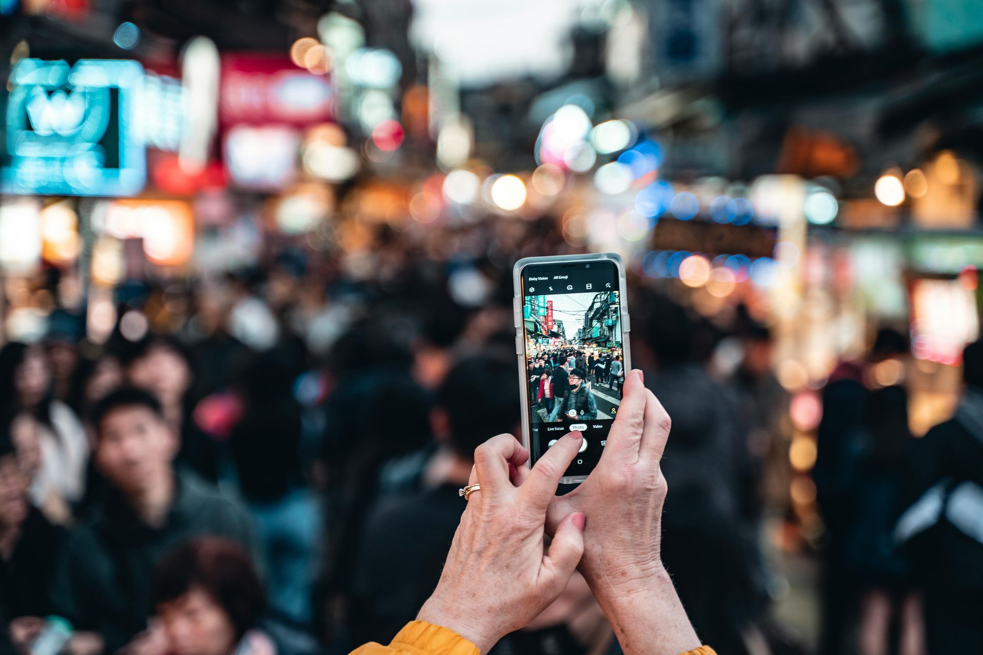 Hands holding a phone, taking a photo of a crowded street with colorful signs and blurred people in the background.
