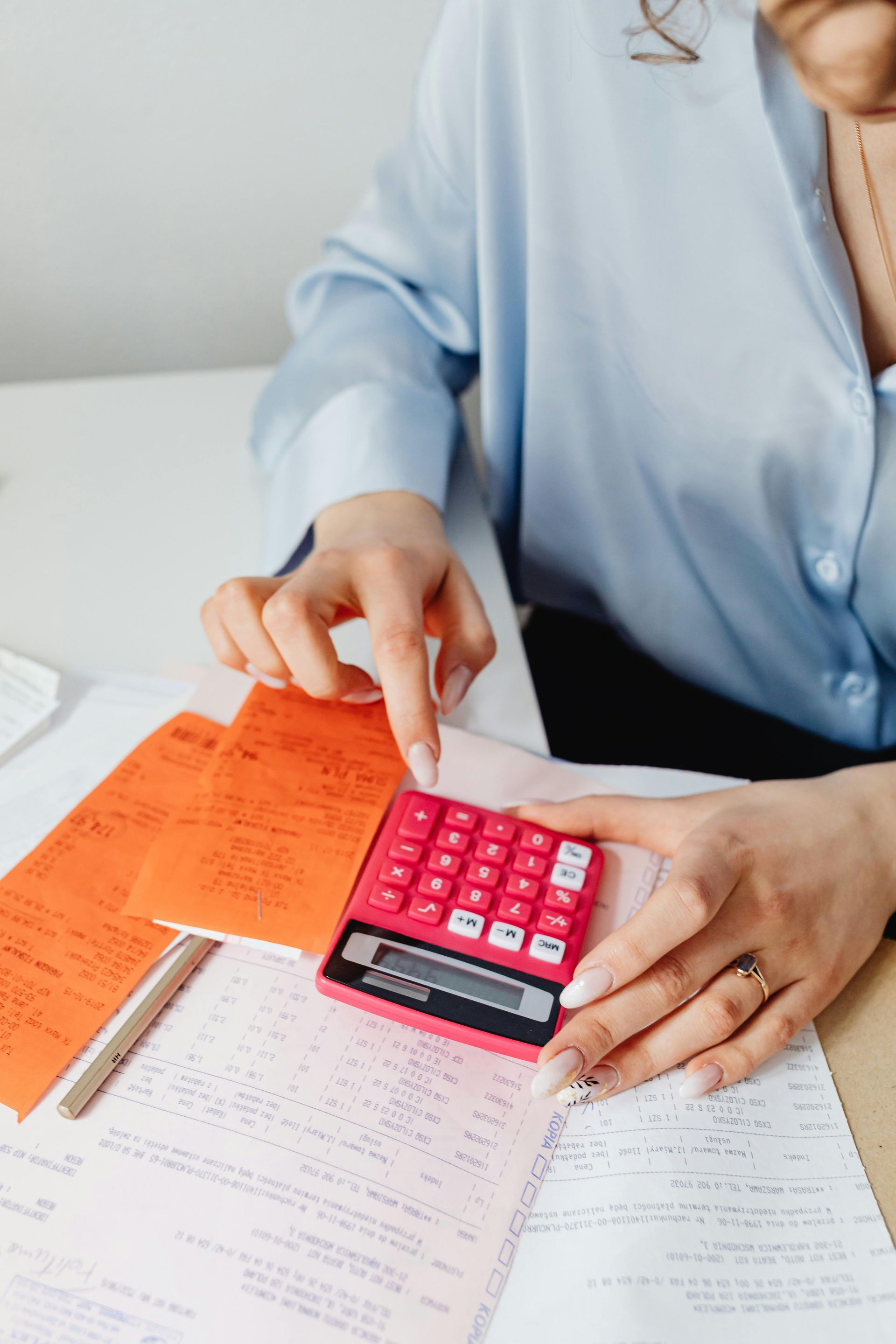 Woman using a pink calculator with financial papers on a white desk; orange folder is open.