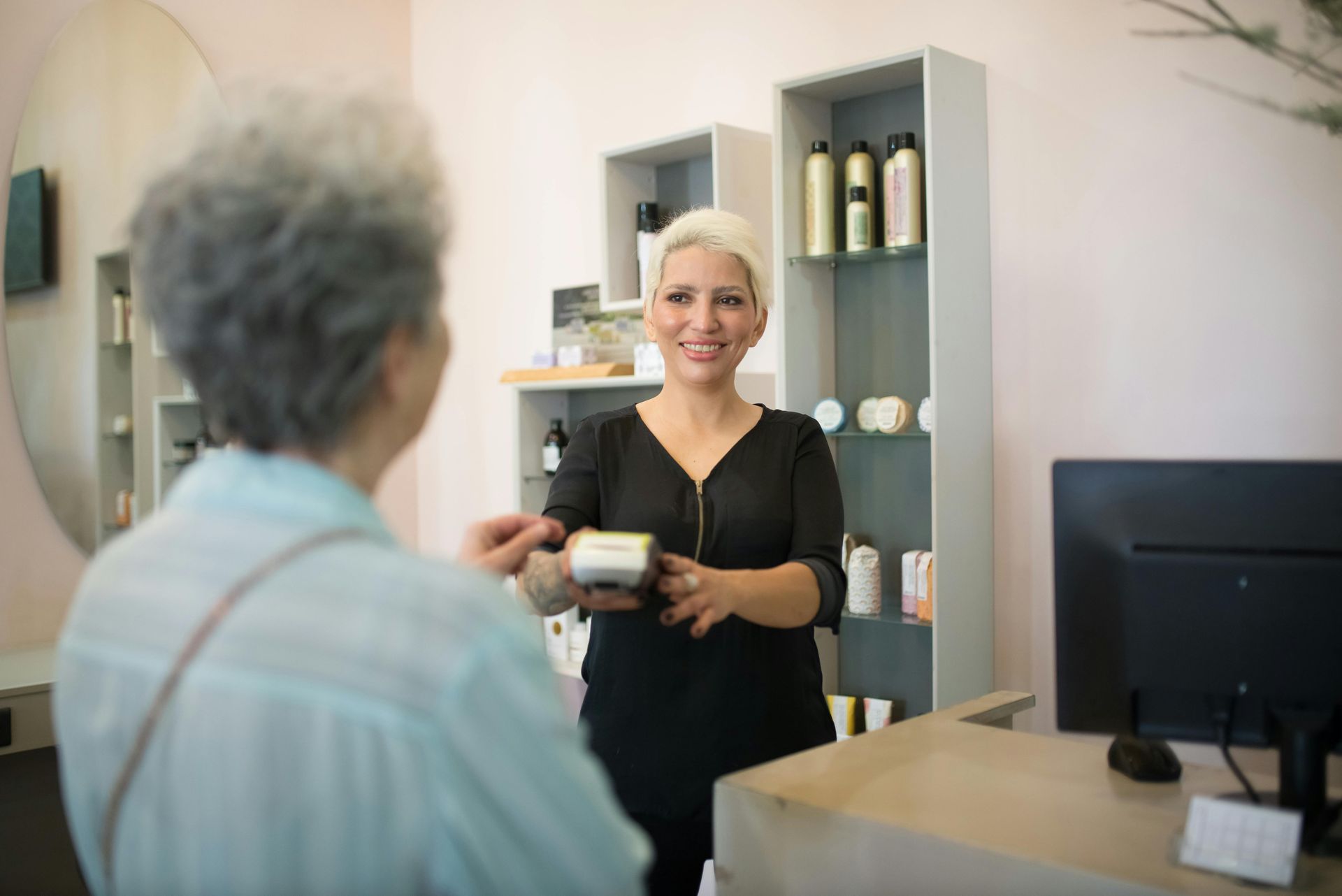 Woman paying at a salon checkout. Smiling salon employee hands her a product, shelves with products in background.
