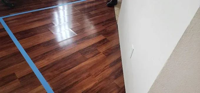Shiny wooden floor with blue tape, a white wall, and a glimpse of a person's legs.