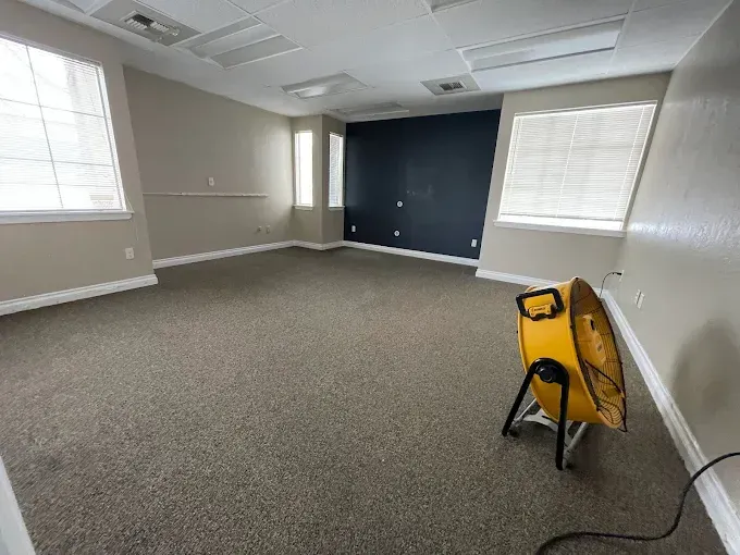 Empty office with carpet, windows, and a fan. A dark blue accent wall is present.
