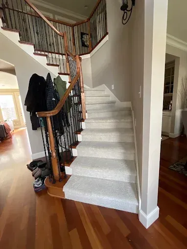 Staircase with carpeted steps, wooden handrail, and black iron spindles. Coats hang on a nearby rack.