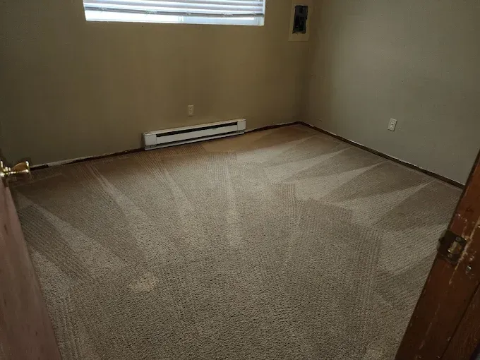 Empty room with beige walls, patterned brown carpet, baseboard heater, and closed door.