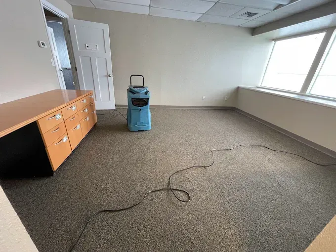 An empty office room with a dehumidifier on the carpet floor near a window. A desk is on the left.