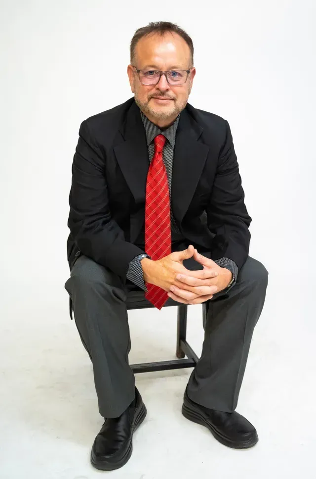 Man in a black suit with a red tie sits on a stool, hands clasped, smiling. White background.