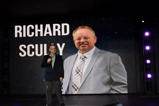 Man standing on stage, holding a microphone. Large screen behind displays the name “Richard Sculley” and his headshot.