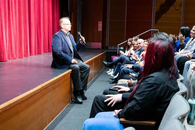 Man speaking on stage to an audience in an auditorium. He is holding a microphone and the red curtain is behind him.