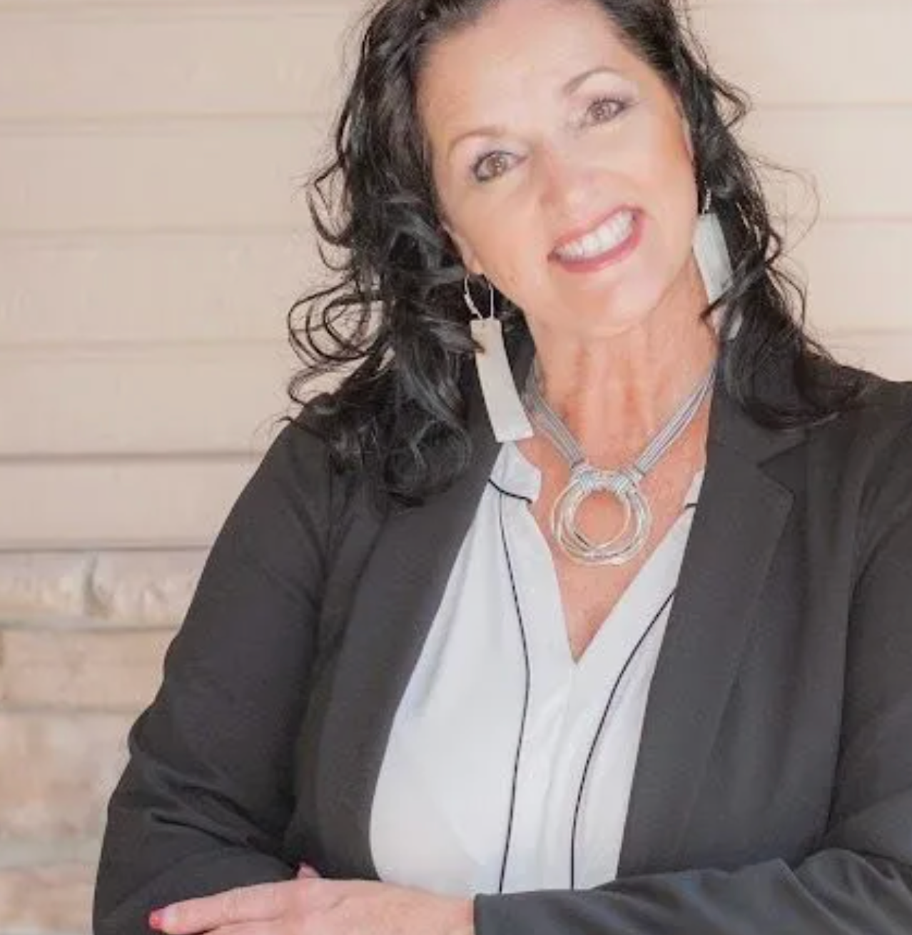 Woman smiling, wearing a black blazer, white blouse, and silver necklace. Leaning against a beige wall.