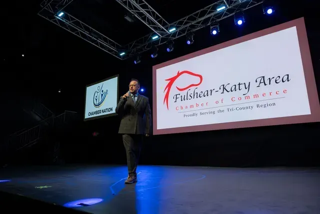 Man on stage speaking at a Chamber of Commerce event, Fulshear-Katy Area logo displayed.