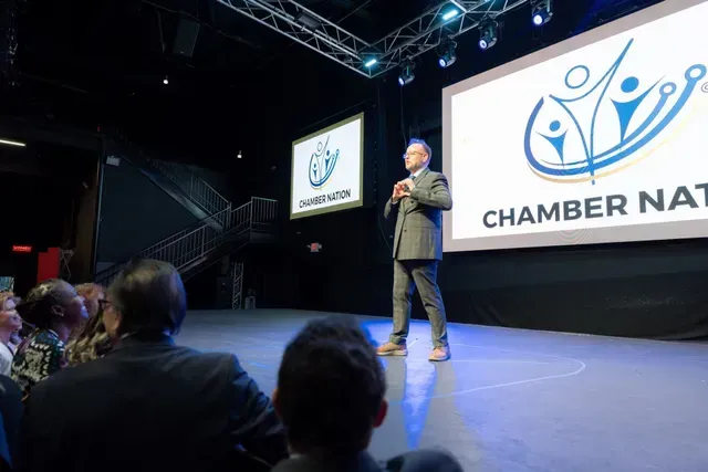 Man presenting on stage, Chamber Nation logo in background, audience seated.