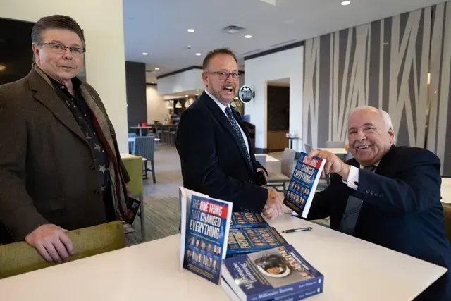 Three men at a table, signing books. One holds a book, smiling. Other books are on the table. Neutral setting.