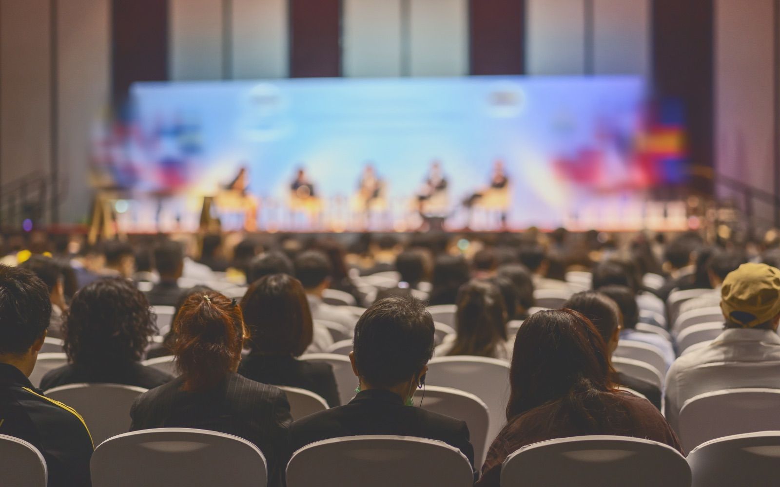 Audience watching a panel discussion on a stage at a conference. The stage backdrop is colorful.