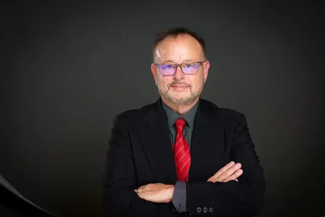 Man with glasses and beard in suit with red tie, arms crossed, smiling.