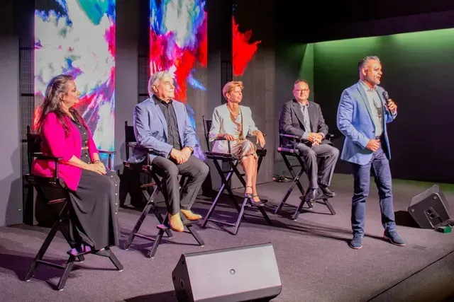 Panel of five people on stage; one speaking with microphone. Abstract background with color lights.