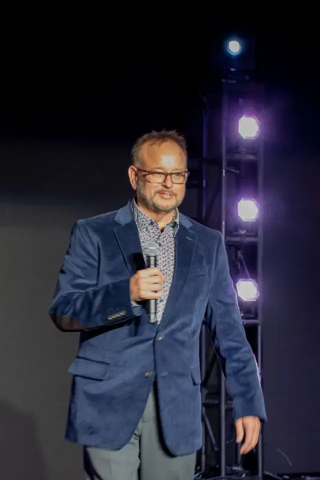 Man in blue velvet jacket, holding a microphone, on a stage with lights.