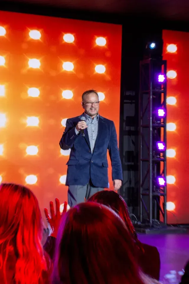 Man in blue blazer speaks on stage with orange and purple lights. Audience in foreground.