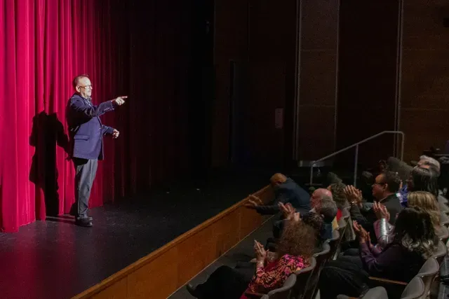 Man on stage points at the audience, who are applauding. Red curtain backdrop, theater setting.